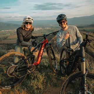Two mountain bikers with helmets and e-bikes posing on a scenic hilltop trail at sunset.