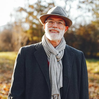 Smiling senior man wearing a fedora hat and grey scarf in a sunny autumn park.