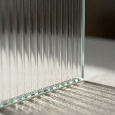 Close-up of light filtering through a ribbed glass panel, casting soft linear shadows on a pale linen surface.