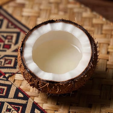Close-up of a half-coconut shell filled with pale kava liquid, placed on woven pandanus mat, tungsten warm light from the left, deep shadow on the right, Fijian textile pattern partially visible beneath the shell