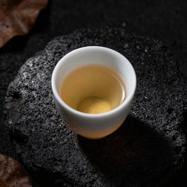 Overhead close-up of an awa cup on dark volcanic stone, pale amber liquid catching diffused window light from the left, dried leaves visible at the edge of frame, moody low-contrast Hawaiian aesthetic