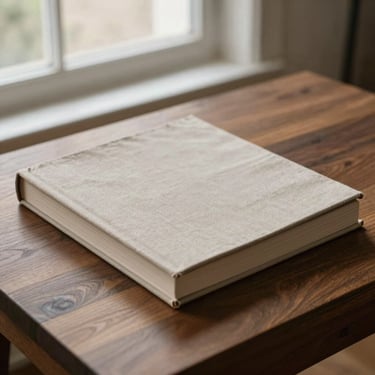 A beautifully bound linen art monograph resting on a dark walnut table, soft morning daylight from a nearby window, quiet studio atmosphere.