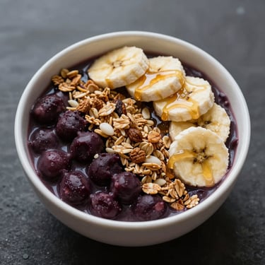 Overhead close-up of a dark purple açaí bowl topped with granola, banana slices and honey drizzle in a white bowl, soft studio light