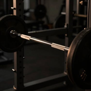 Dramatic low-key side-lighting on a barbell being lifted from a rack, metallic surfaces reflecting warm gold highlights, dark charcoal tones.