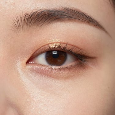 Macro close-up, soft diffused morning light. A detailed view of luminous, hydrated skin texture showing real pores, with a subtle touch of gold shimmer on the eyelid and clean, defined lashes.
