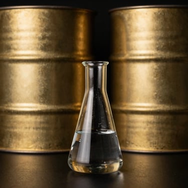 Clean laboratory glassware with a dark liquid, industrial stainless steel barrels in the background, brushed metallic gold details, high-contrast lighting