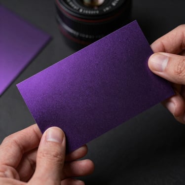 Macro shot of physical brand collateral with metallic violet foil, low-key lighting, hands holding a premium dark paper card.