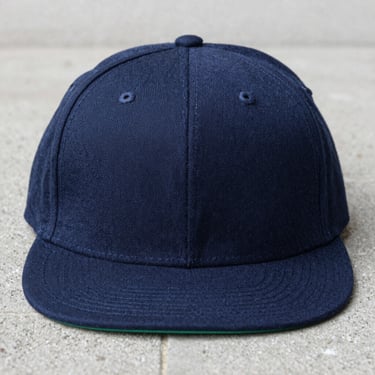 Front-facing product shot of a classic Yupoong flat-bill snapback cap in navy blue, concrete background, sharp focus, 50mm lens