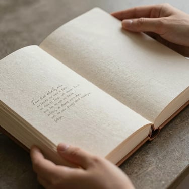 Macro shot of hands gently holding an open linen memory album under soft, diffused morning light, showing thick textured paper and quietly personalized embossed typography.