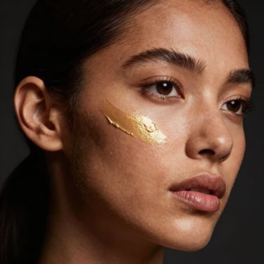 High-angle dramatic portrait of a model with deep skin tone, sharp spotlight highlighting pure gold liquid illuminator on the cheekbones, dark background.