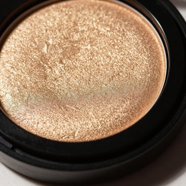 Luminous gold highlighter powder pressed into a sleek black compact, sharp side lighting highlighting the texture