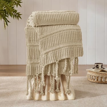 Cream chunky knit throw blanket with tassels draped over a stool in a cozy living room.