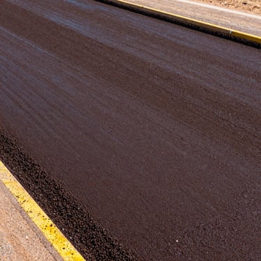 Technical close-up of dark brown cationic bitumen emulsion being applied to a road base, showing fine liquid texture