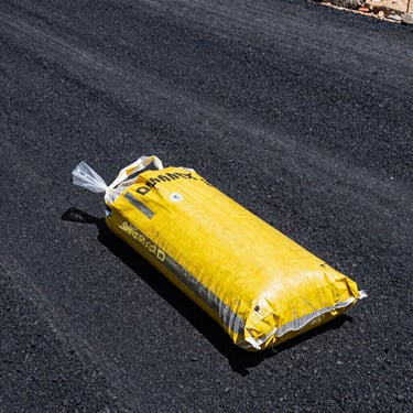 A heavy plastic bag of premium cold mix asphalt sitting on a freshly repaired road surface, showing coarse black aggregate