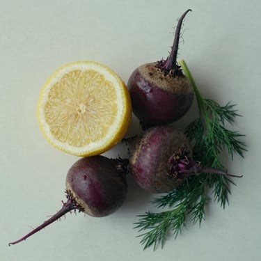 Overhead close study of mixed seasonal produce — a halved lemon, two small beets, a sprig of dill — arranged on a pale off-white surface in cool north-facing daylight, no props, no garnish