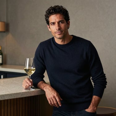 A South American man in a dark navy sweater leaning against a minimalist bar counter, holding a glass of white wine, refined and calm expression.
