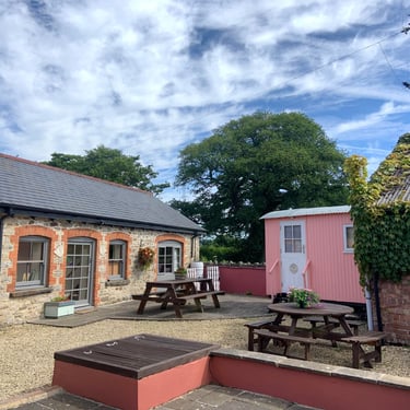Rustic farm courtyard with a stone cottage, pink shepherd hut, and wooden picnic tables under a blue sky.