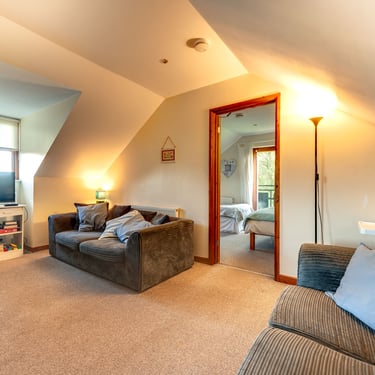 Cozy attic living room with grey corduroy sofas, beige carpet, and a view into a bright bedroom.