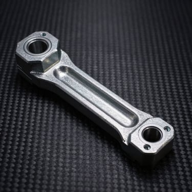 Heavy-duty steel suspension control arm, isolated on dark carbon surface, high-contrast side-lighting.