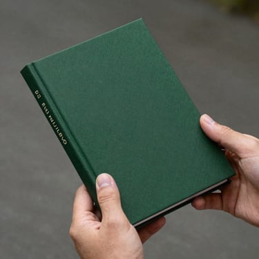 A hand holding a slim, hand-bound photobook with a dark green fabric cover and minimalist cream typography on the spine