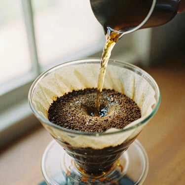 Close-up of water pouring over coffee grounds during the bloom phase in a glass dripper, warm amber tones, soft side-lighting, 35mm film.
