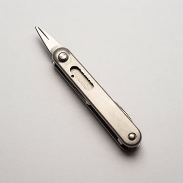 Top-down flat-lay of a sleek titanium multi-tool pen on a clean off-white background, soft studio lighting