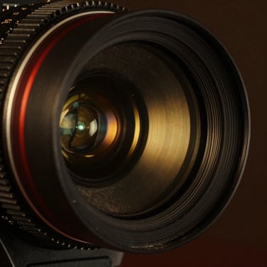 Extreme close-up of an anamorphic cinema lens, reflection of a golden stage light in the glass, deep shadows.