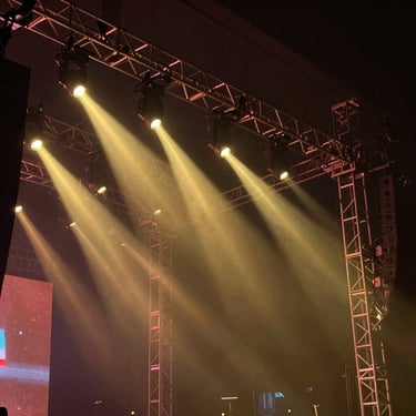 Low-angle shot of a custom concert stage truss, dramatic golden spotlights piercing through haze.