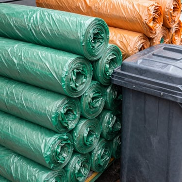Roll of heavy-duty garbage bags and a portable trash can next to a market stall, emphasizing cleanliness, overcast daylight.