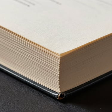 Close-up of a physical book showing heavy paper texture, exposed thread binding, high-contrast studio lighting, sharp shadows.