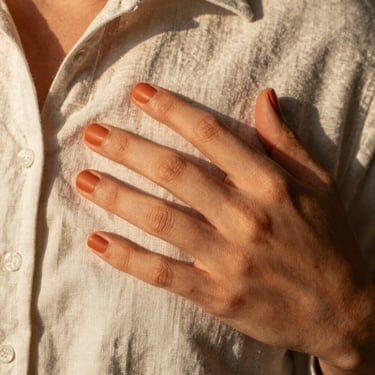 Close-up of hand with terracotta nails resting on a linen shirt, warm natural daylight