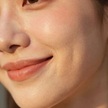 Macro shot of a smiling woman's cheek showing natural skin texture and a soft peach blush, warm golden hour lighting.