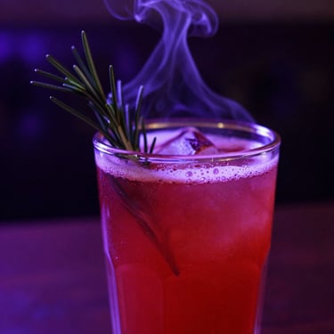A vibrant crimson cocktail garnished with a smoking rosemary sprig, low-key neon purple lighting, macro shot.