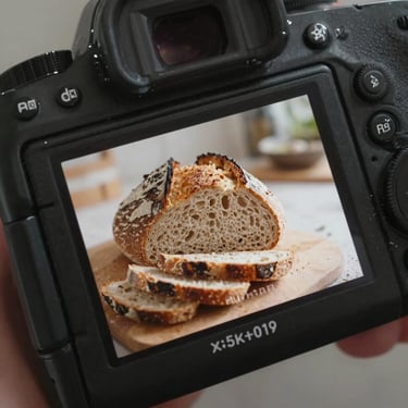 Close-up of a camera screen displaying a live shoot of fresh sourdough bread being sliced, warm morning light, hands of the photographer visible.