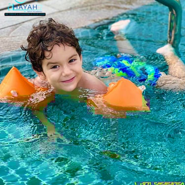 Smiling young boy wearing orange arm floaties during a swimming lesson in a turquoise pool.