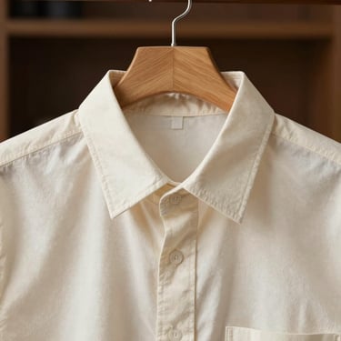 Close-up of a cream white cotton-blend shirt hanging on a raw wooden dowel, detailing the subtle roll of the collar
