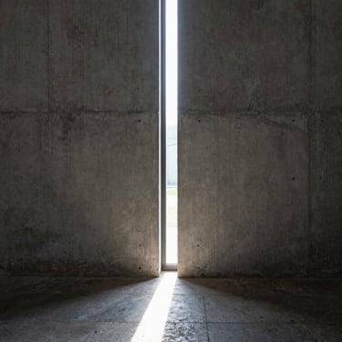 A narrow vertical window slicing through a thick concrete wall, casting a sharp beam of light on a stone floor.