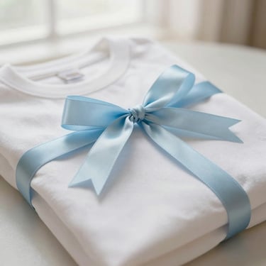 Close-up of a coquette-inspired personalized tee neatly folded with a pastel blue ribbon tied around it, soft morning light, shallow depth of field