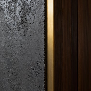 Close-up architectural detail of premium structural materials, textured dark concrete meeting brushed brass and dark wood paneling, dramatic side-lighting showing texture, professional architectural photography.