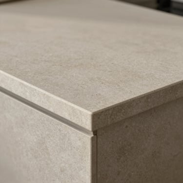 Close-up of stone-look countertop surface, light grey-beige tone, subtle texture under warm ambient light, edge detail and corner visible, no items on surface
