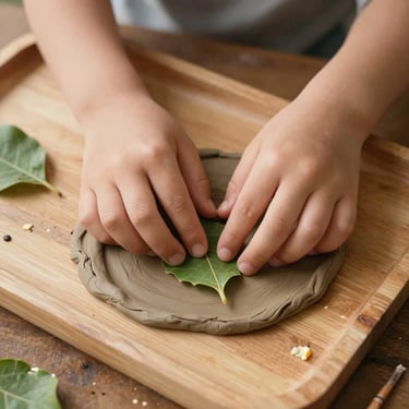Overhead close-up of a child's hands pressing a leaf into soft clay on a wooden tray, natural afternoon light, scattered petals and a small twig visible at the edges