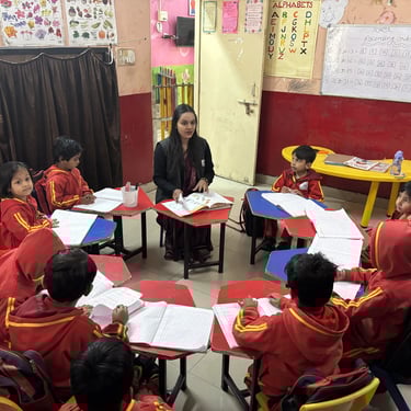 A primary school teacher sits with students in a circle for a lesson in a colorful classroom.
