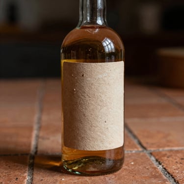 A heavy glass bottle with an earthy clay-textured label, placed on baked clay tiles, moody low-key lighting.