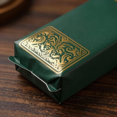 A close-up of a premium dark green package with intricate gold foil patterns, resting on a polished dark wood surface.