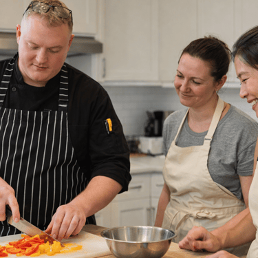 A chef conducts a cooking class for two clients in a home kitchen.
