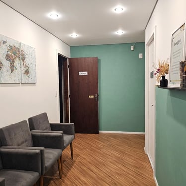 Sala de espera com cadeiras cinza, quadros na parede, portas com sinalizações e prateleira com enfeites.