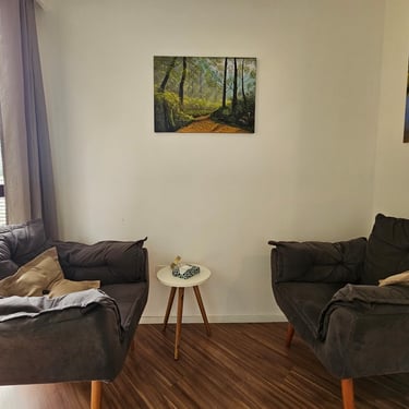 Canto de uma sala com duas poltronas cinza escuro, mesa branca redonda com item decorativo, quadros pendurados nas paredes.