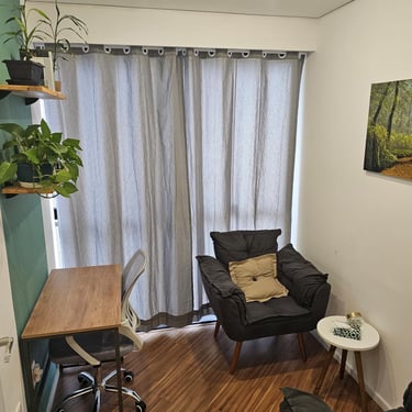 Consultório com cadeira de escritório, mesa de madeira, poltronas cinzas com almofadas, cortina e plantas.