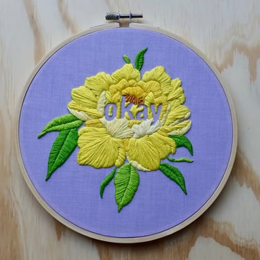 A large yellow embroidered flower on purple cotton with green leaves and the word "okay" against a wooden background