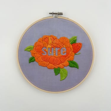 Orange embroidered 2-D flower on purple fabric against white background. The word "sure" is stitched in the negative